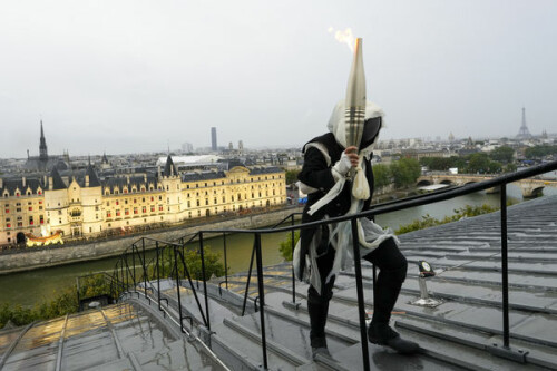 A torchbearer carries the Olympic flame over a building along the Seine River in Paris, France, during the opening ceremony for the 2024 Summer Olympics, Friday, July 26, 2024. (AP Photo/Bernat Armangue, Pool)